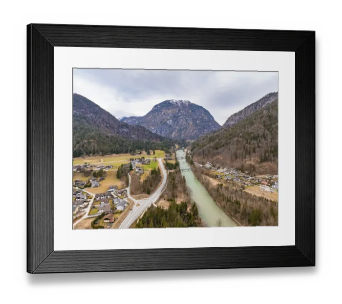 Framed Print : Aerial view of a rural area with a river flowing past a settlement and surrounded by mountains under a cloudy sky
