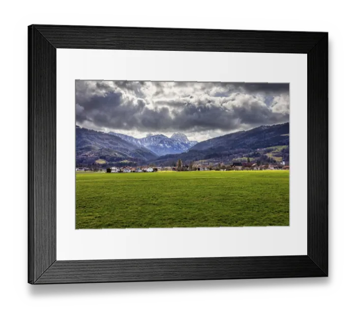 Framed Print : Dramatic Alpine landscape with green meadow in the foreground and cloudy sky over a village Framed Print : Dramatic Alpine landscape with green meadow in the foreground and cloudy sky over a village