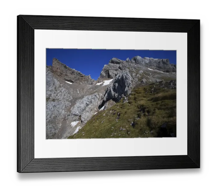 Framed Print : Steep mountain face with scattered snow fields and green grassy areas under a clear sky