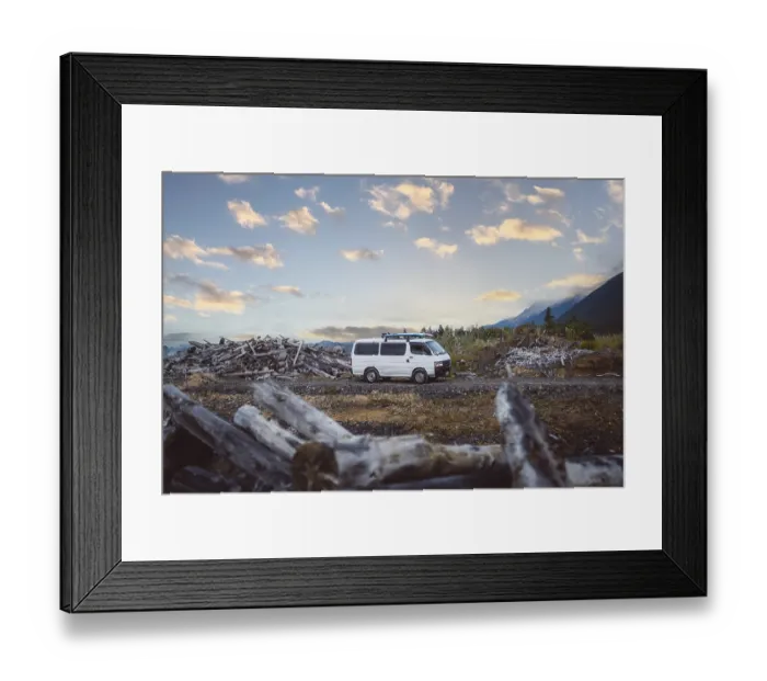 Framed Print : A van stands in a rural area between piles of wood and mountains in the background