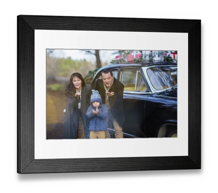 Framed Print : Smiling adult man and woman with boy in warm clothes blowing snow standing at vintage car with Christmas presents
