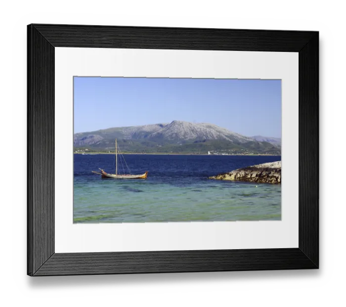 Framed Print : A wooden boat floats on a clear blue sea in front of a large mountain Framed Print : A wooden boat floats on a clear blue sea in front of a large mountain