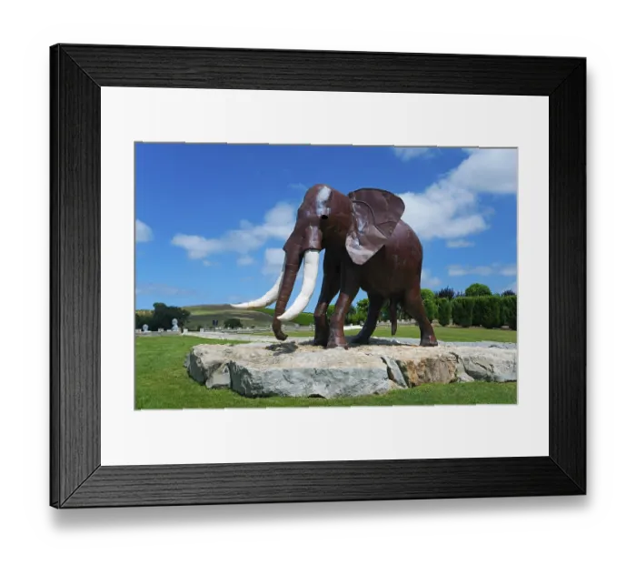 Framed Print : Metal sculpture of an elephant on a rock foundation in a green park under a blue sky with white clouds