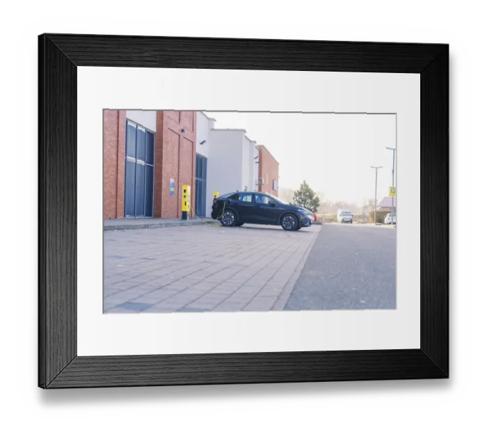 Framed Print : An electric car charging in a car park in front of a modern building in daylight Framed Print : An electric car charging in a car park in front of a modern building in daylight