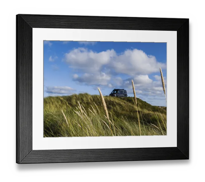 Framed Print : A campervan stands on a grassy dune under a blue sky with clouds