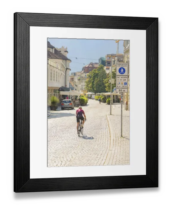 Framed Print : A cyclist rides along a sunny city street surrounded by historic buildings and trees