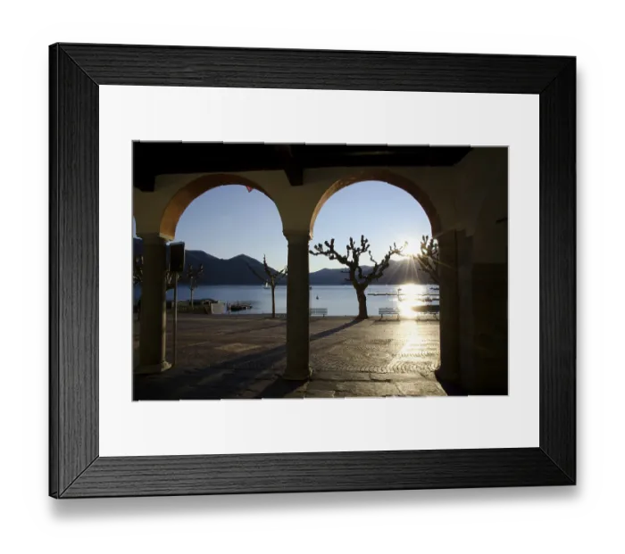 Framed Print : Arch and Bare Tree on the Waterfront to Lake Maggiore with Mountain and Sunbeam in Sunset in Ascona