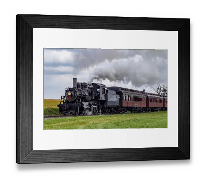 Framed Print : A vintage steam train chugs along railway tracks surrounded by lush green fields under a clear blue sky