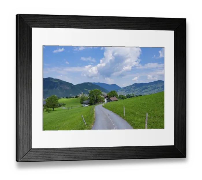 Framed Print : Country road leads through green meadows into a rural mountain landscape under a blue sky with white clouds