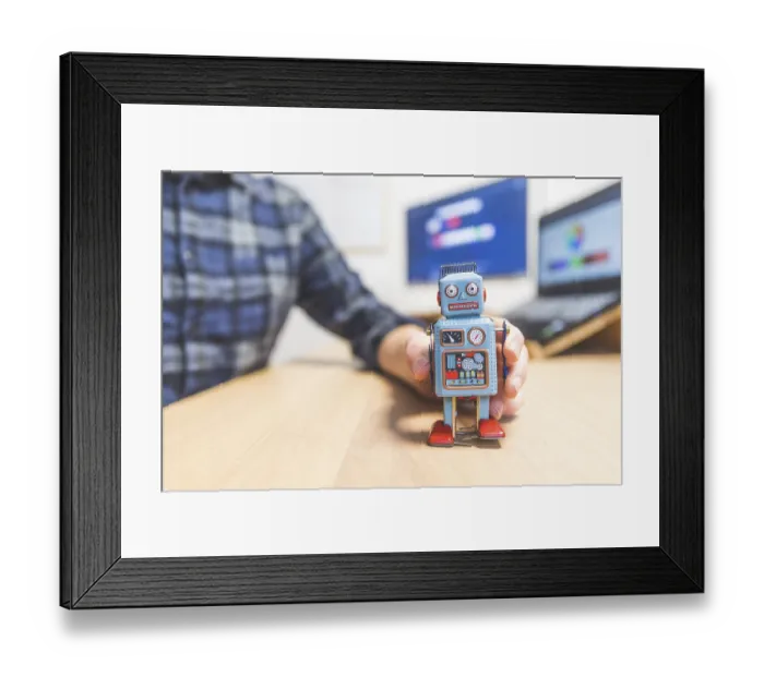 Framed Print : Toy robot on a desktop, man working on the computer in the background Framed Print : Toy robot on a desktop, man working on the computer in the background