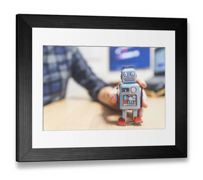 Framed Print : Toy robot on a desktop, man working on the computer in the background Framed Print : Toy robot on a desktop, man working on the computer in the background