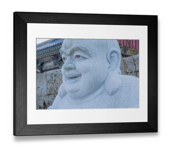 Framed Print : Closeup of head and face of stone carved sitting Buddha with blurred temple building in background in Pyeongtaek