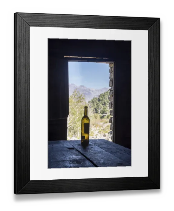 Framed Print : An empty wine bottle on a wooden table in front of an open door with a view of the mountains