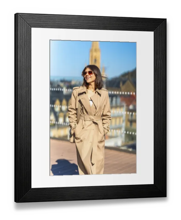 Framed Print : A woman wearing a tan trench coat is smiling and walking on a bridge