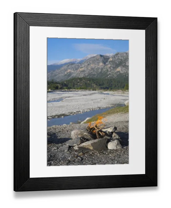 Framed Print : A campfire in a rocky landscape with mountains and a riverbed in the background