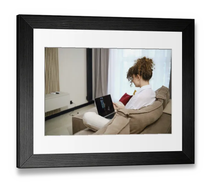 Framed Print : A young woman sits on the couch in the living room and reviews her photos on a laptop