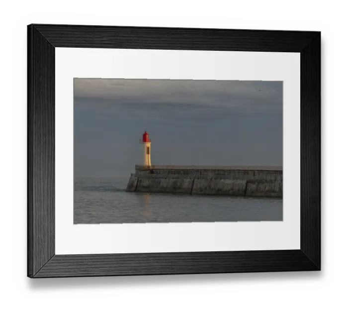 Framed Print : A lighthouse stands on a pier and casts its light over calm waters at dusk