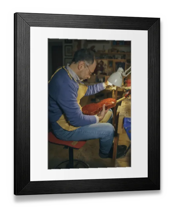 Framed Print : Violin maker inspecting the instrument under the light in his workshop crafting a stringed instrument using traditional