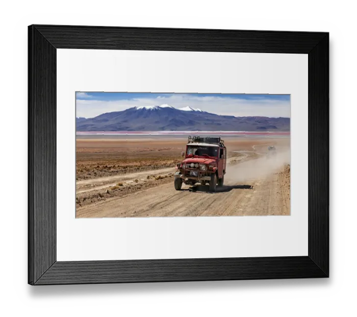 Framed Print : A jeep drives dusty on a desert road with mountains in the background