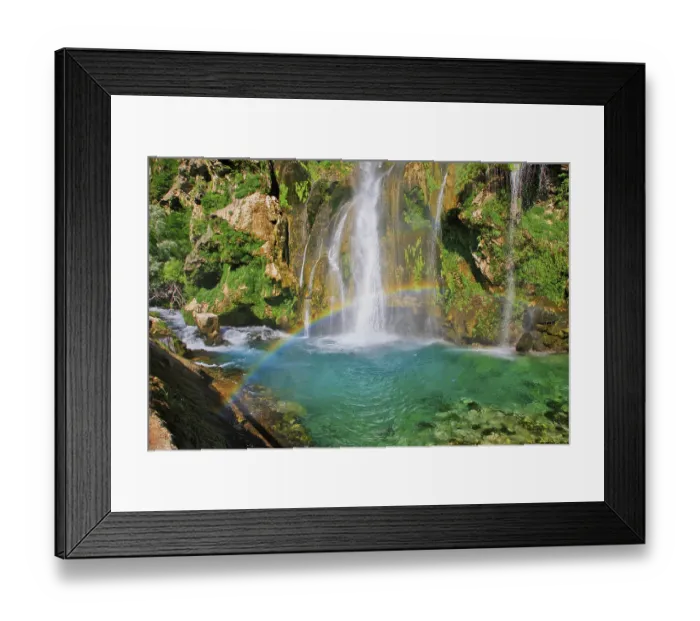 Framed Print : Waterfall with rainbow, surrounded by rocks and green foliage under a clear sky