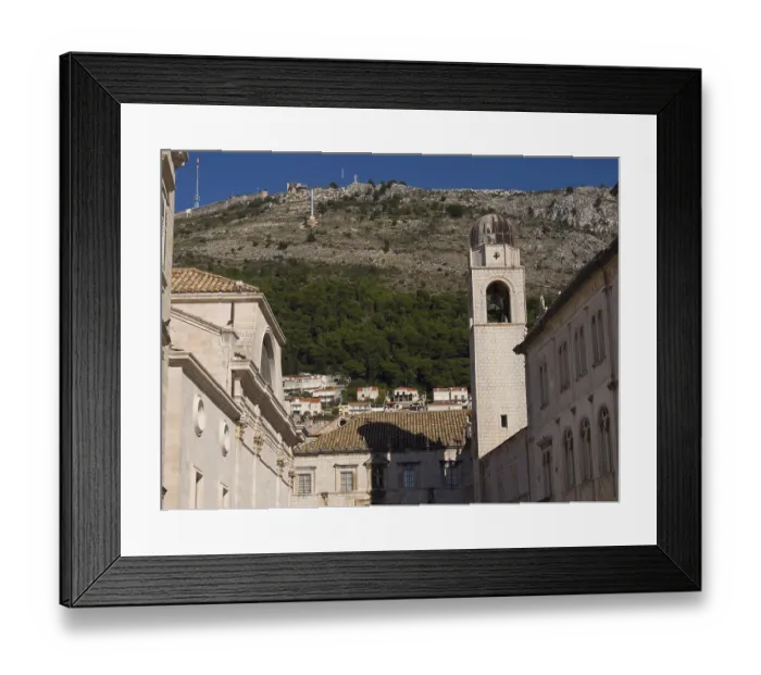 Framed Print : Church bell tower and historic buildings in front of a wooded mountain and blue sky