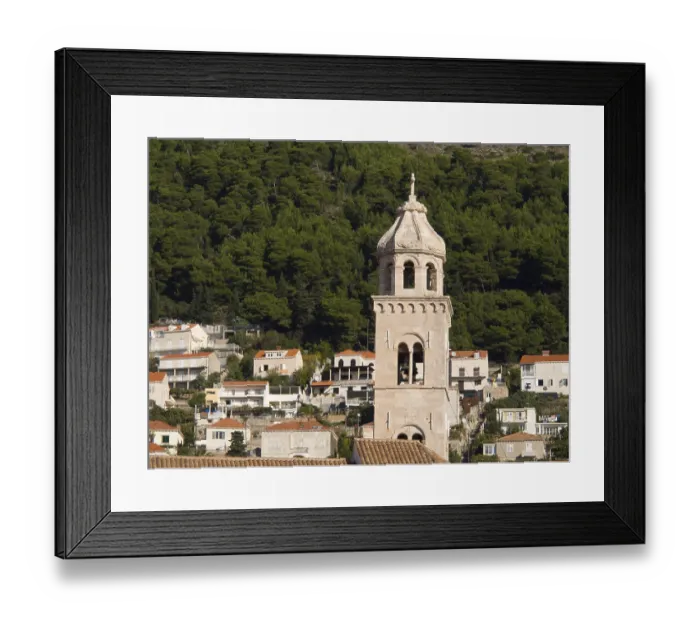 Framed Print : Historic bell tower surrounded by houses and green vegetation in front of a densely wooded hill