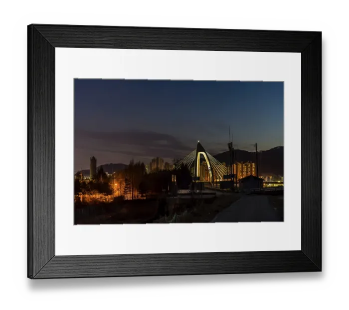 Framed Print : Night view of concrete bridge with triangular arch over a river with community buildings in background