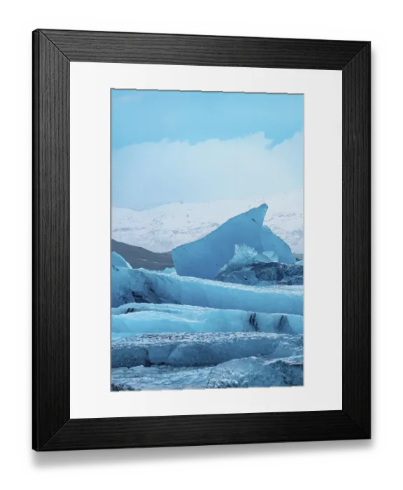 Framed Print : A stark blue iceberg rises against a cold wintry sky with snowy peaks in the distance