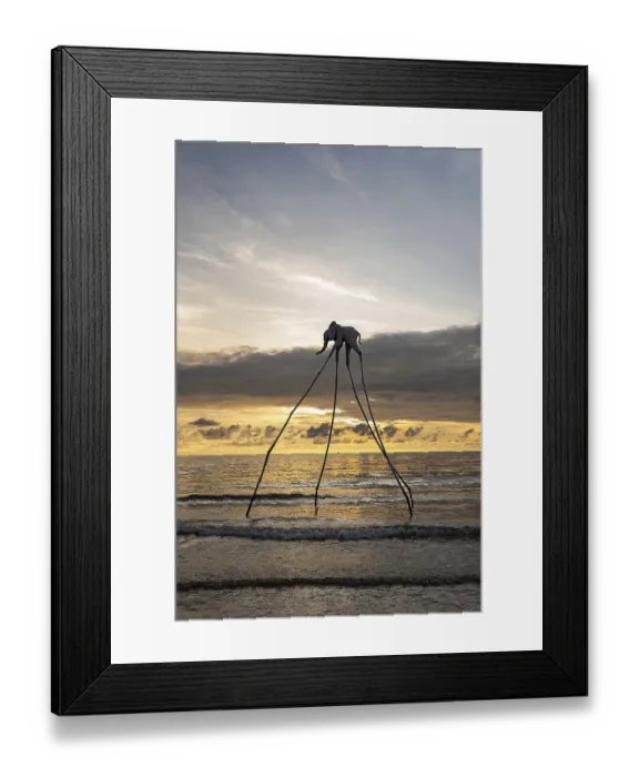 Framed Print : Surreal elephant sculpture with long legs by the ocean at sunset with gentle waves