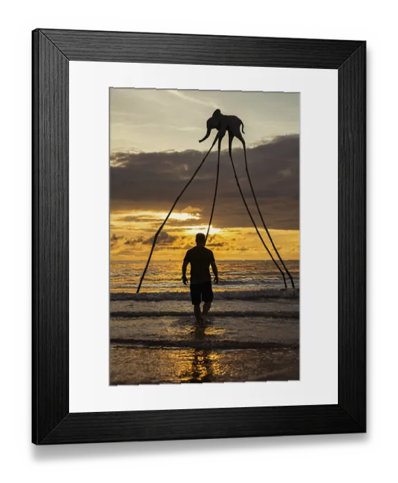 Framed Print : A man stands in the surf facing a surreal elephant sculpture with long legs