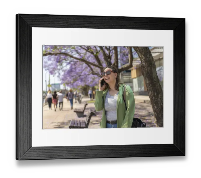 Framed Print : A cheerful young woman with sunglasses and a green jacket talks on her phone during a sunny day on the riverfront