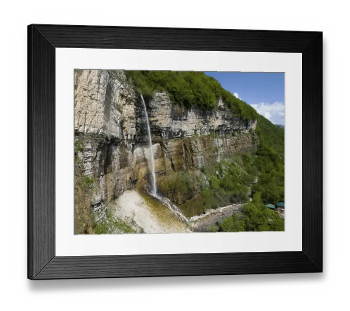 Framed Print : Long waterfall falls over a rock face into a wooded area under a clear sky