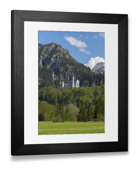 Framed Print : Neuschwanstein Castle towers over a green meadow with a dramatic mountain backdrop under a blue sky Framed Print : Neuschwanstein Castle towers over a green meadow with a dramatic mountain backdrop under a blue sky