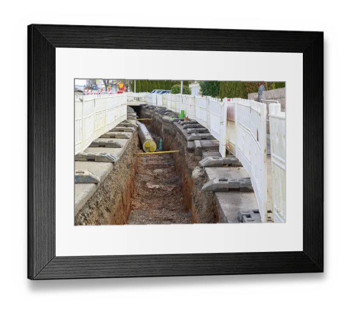 Framed Print : Construction site scene with district heating pipeline in a deep trench and white barriers