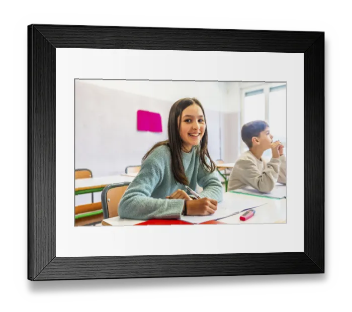Framed Print : Happy schoolgirl writing on notebook during lesson in classroom with schoolmate thinking in background