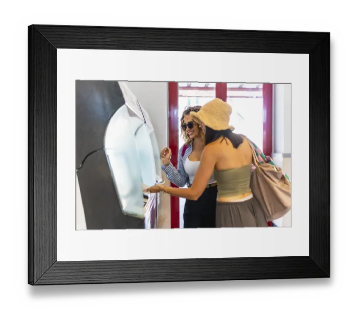 Framed Print : Two happy female tourists buying train tickets at automatic ticket machine inside station building