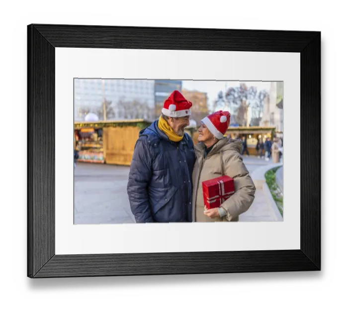 Framed Print : Loving senior couple wearing santa hats exchanging christmas gifts at a christmas market in the city