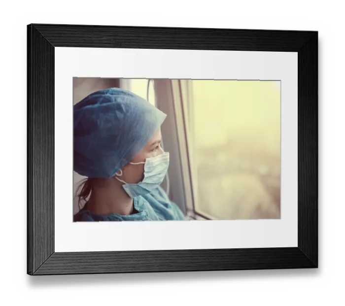 Framed Print : A healthcare worker in scrubs and a mask gazes out the window at sunset