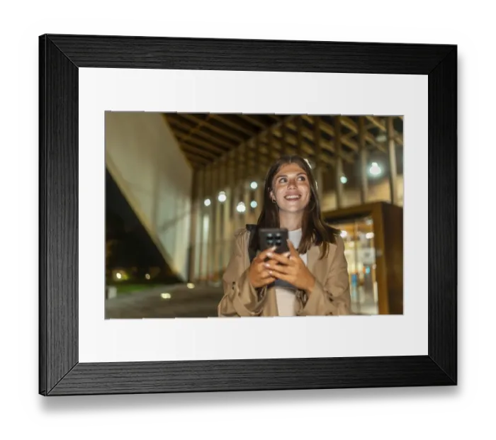 Framed Print : Young woman smiling while holding a smartphone and looking up