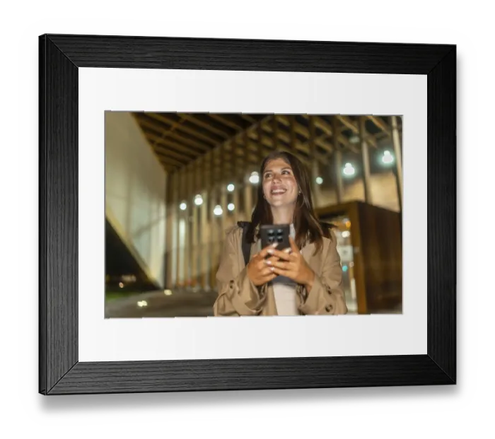 Framed Print : Young woman smiling, holding phone while walking illuminated city streets at night