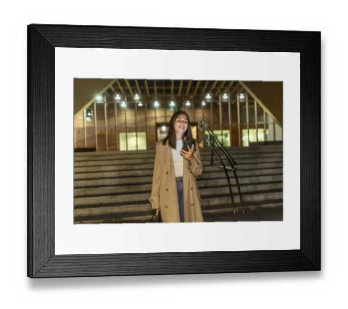 Framed Print : Young woman in a trench coat smiling and looking at her smartphone while standing on modern urban stairs at night