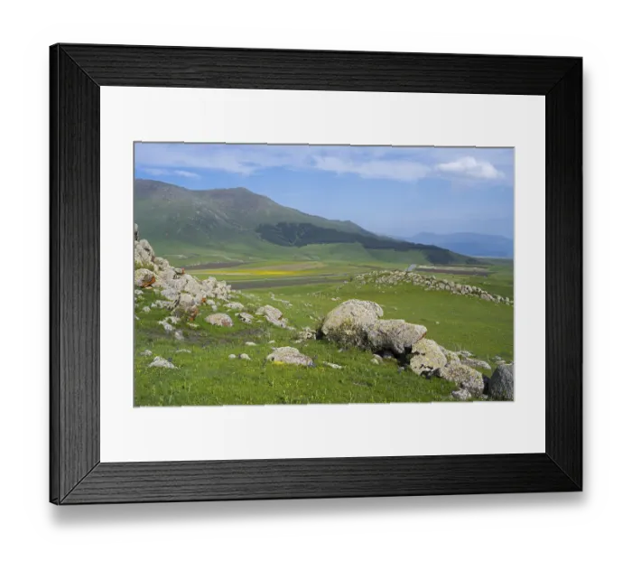 Framed Print : Green hilly landscape with rocks and mountains in the background under a slightly cloudy sky Framed Print : Green hilly landscape with rocks and mountains in the background under a slightly cloudy sky