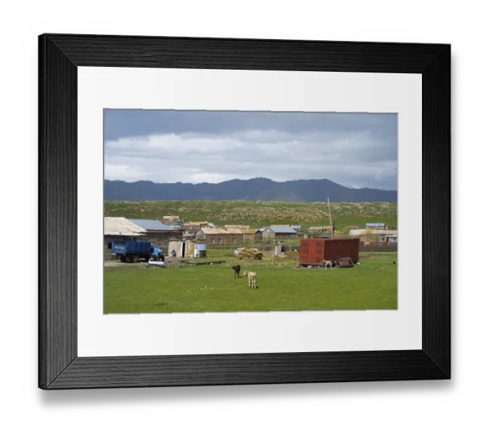 Framed Print : Rural settlement with huts and animals on a green pasture surrounded by mountains under a cloudy sky