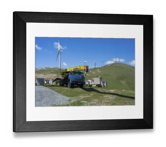 Framed Print : A blue crane lorry stands on a green construction site next to a wind turbine on a hill