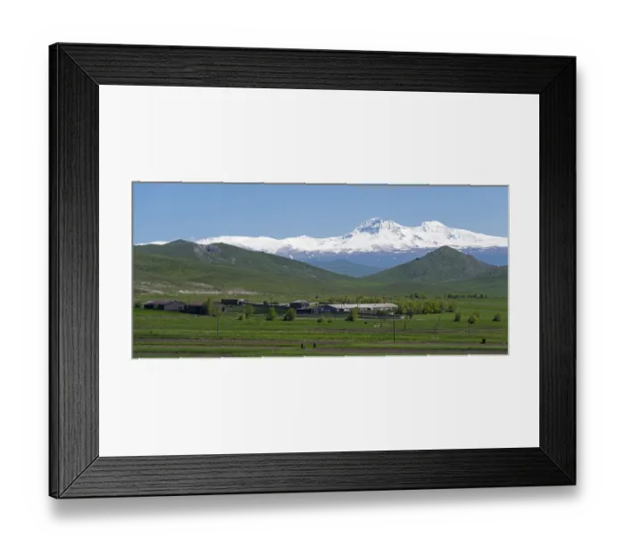 Framed Print : A mountain village lies in the middle of green fields in front of a snow-covered mountain range