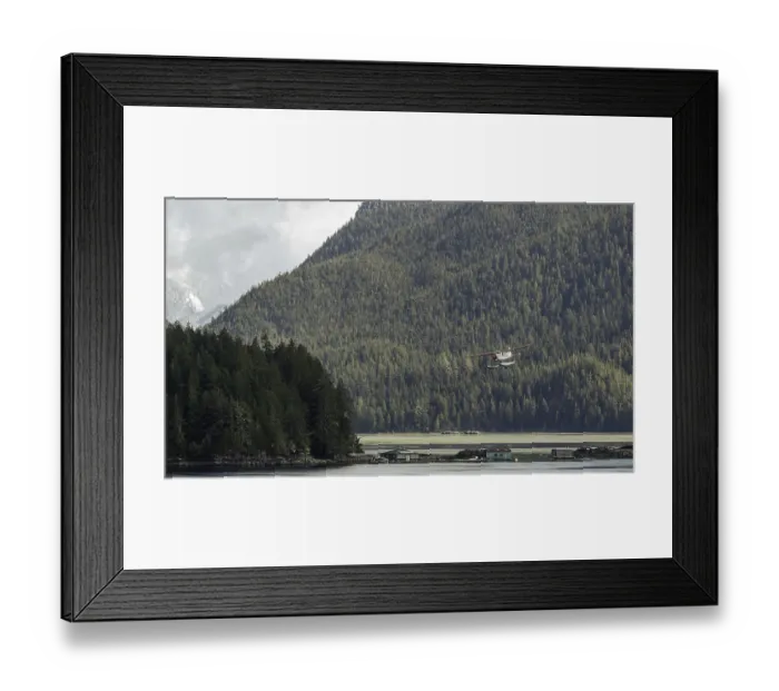 Framed Print : A seaplane flies over a tranquil lake surrounded by dense forest and mountains under an overcast sky in Tofino