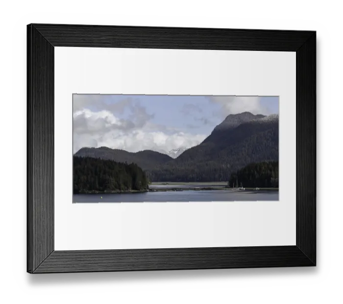 Framed Print : An expansive view of a lake bordered by forests and mountains under a cloudy sky in Tofino