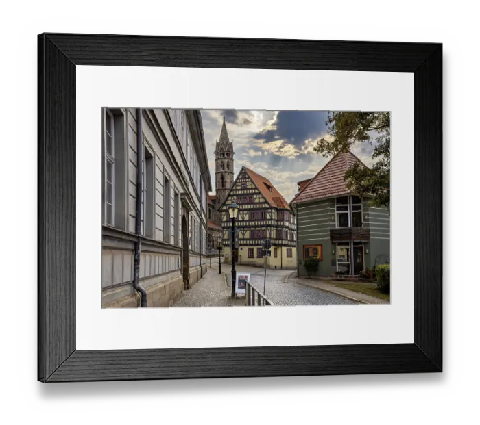Framed Print : Paved street with half-timbered houses and church tower under a cloudy sky