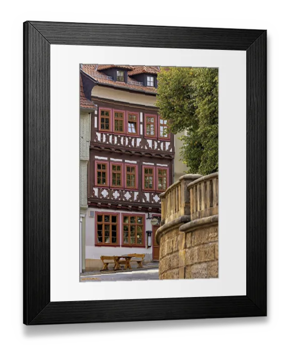 Framed Print : A half-timbered house with red window frames next to a stone wall in a historic town