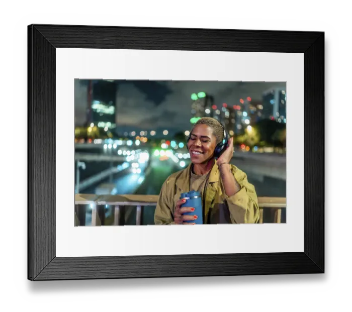 Framed Print : Woman standing on a bridge, holding a travel mug and wearing headphones while happily listening to music alone at night